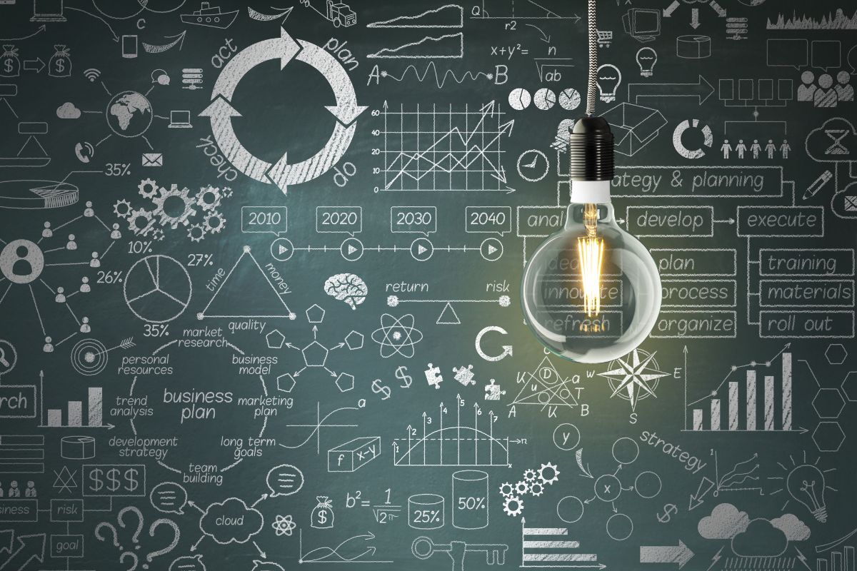 Chalkboard with several technical drawings, graphs and flowcharts. Brightly lit lightbulb hanging in front of the chalkboard.