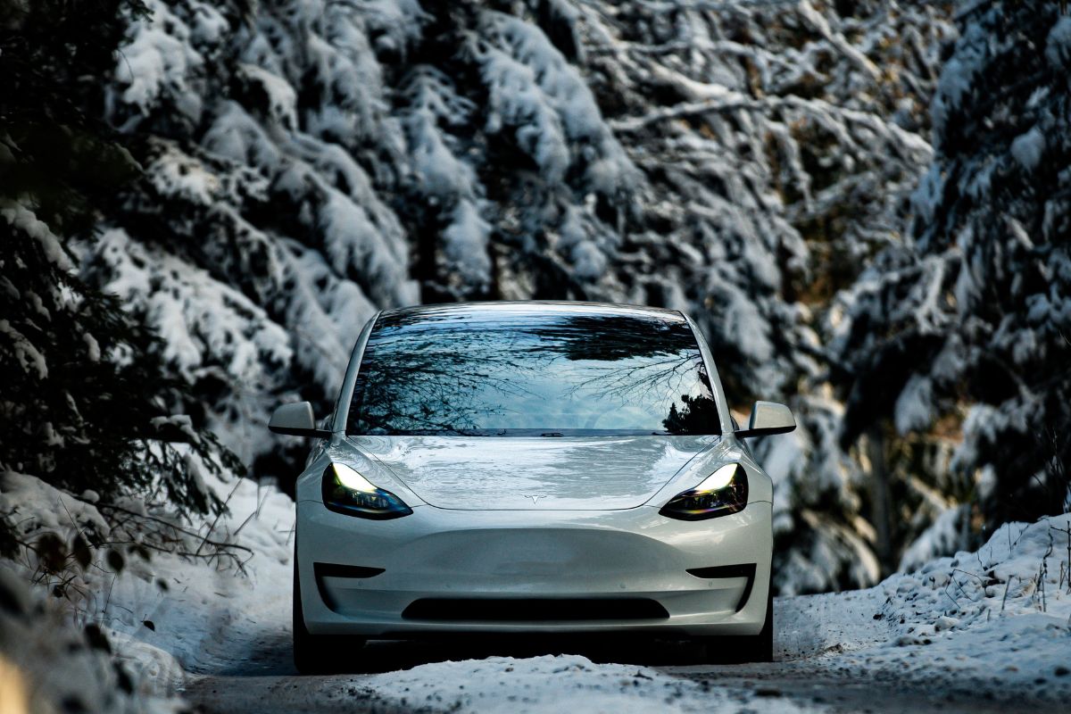 Electric vehicle driving on a snowy road lined with snow-covered pine trees.