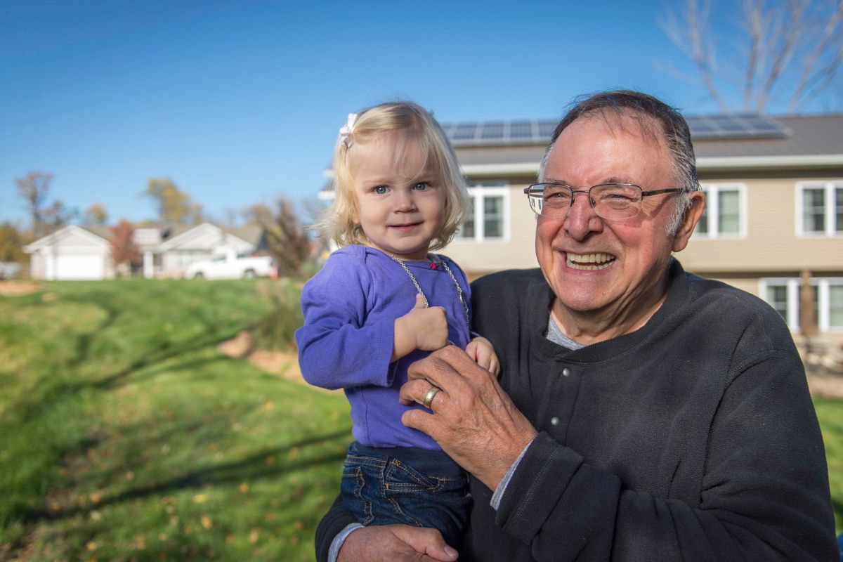 Grandparent holding grandchild, both smiling and standing in front of a house with rooftop solar panels. 