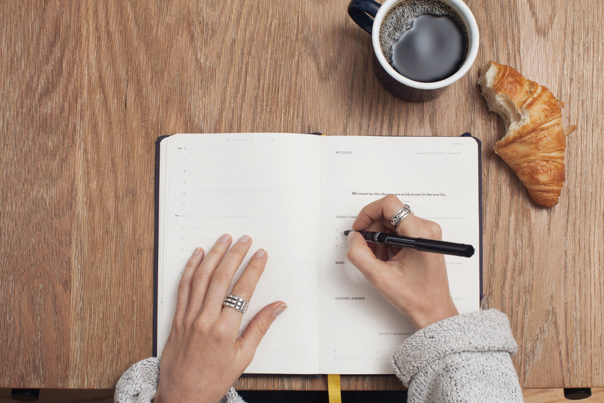 person sitting at a table writing in a planner, enjoying coffee and a pastry