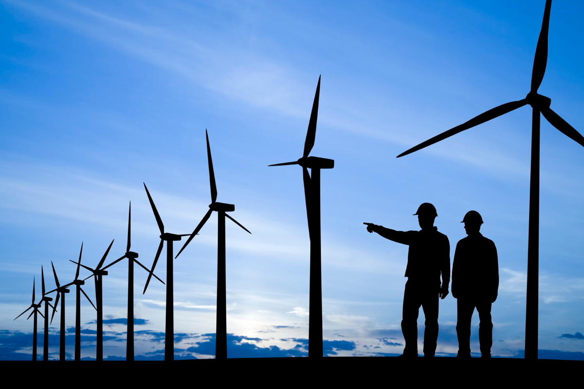 Silhouette of two construction workers standing in an array of wind turbines. The worker on the left is pointing to the left. 