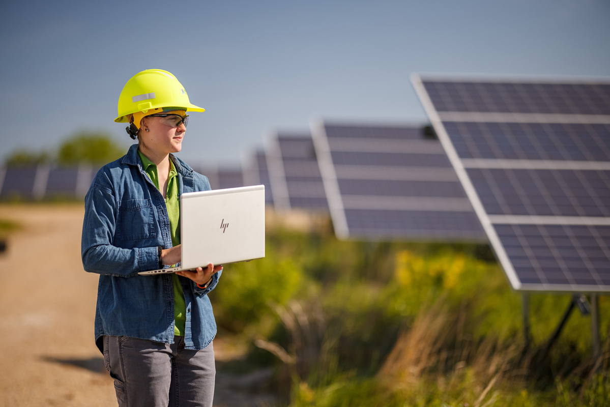 Engineer in hard hat and safety glasses holding a computer looking at rows of solar panels. 