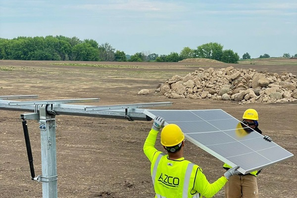 Two workers installing solar panels. A large pile of rocks covers a part of the field behind the workers.
